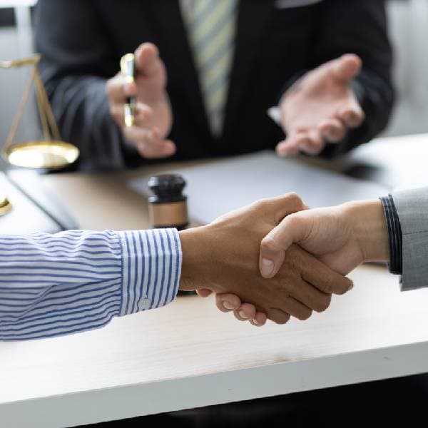 Close-up of two people shaking hands with a lawyer, a gavel, scale, paper, and laptop on white desk in background