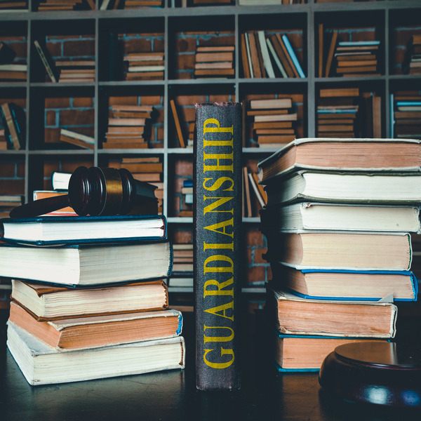 Stacks of books and a gavel on a wooden table with a book standing up in the middle that has “Guardianship” written on the spine
